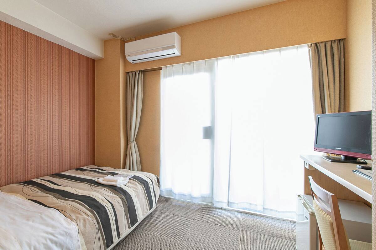 A hotel room with a bed, a desk with a computer, an air conditioner, and a large window with curtains.