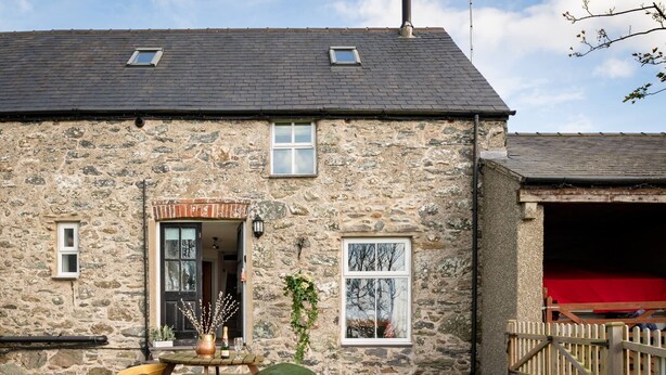 A stone building with a wooden door, a small window, and a picnic table with a cushion. - Hill Top 1: 3-Bed Cottage in Caergeiliog | Managed by Anglesey Holiday Lettings (Caergeiliog)