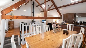 A spacious loft-style room with a wooden dining table, white chairs, and a kitchen area with dark wooden cabinets. - Hill Top 1: 3-Bed Cottage in Caergeiliog | Managed by Anglesey Holiday Lettings (Caergeiliog)