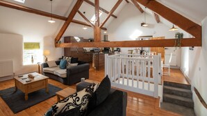 A spacious loft with exposed wooden beams, a living area with a sofa and coffee table, a kitchen, and a dining area with a white partition. - Hill Top 1: 3-Bed Cottage in Caergeiliog | Managed by Anglesey Holiday Lettings (Caergeiliog)