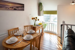 A dining area with a wooden table set for four, a painting on the wall, and a view of the outdoors.