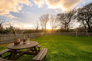 A wooden picnic table with a bottle and glasses, surrounded by a well-maintained lawn and a wooden fence.