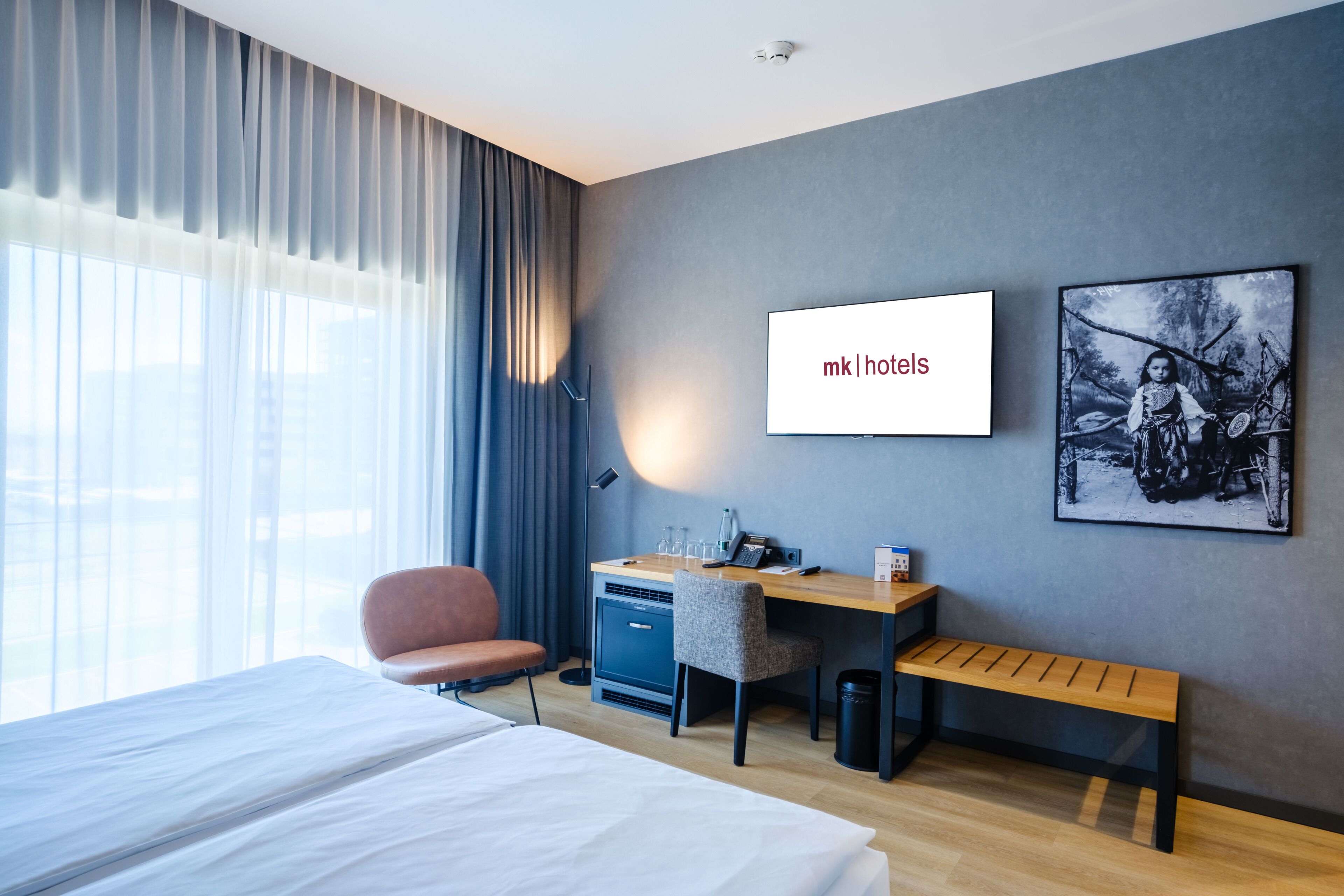 A hotel room with a bed, desk, chair, and a picture on the wall.