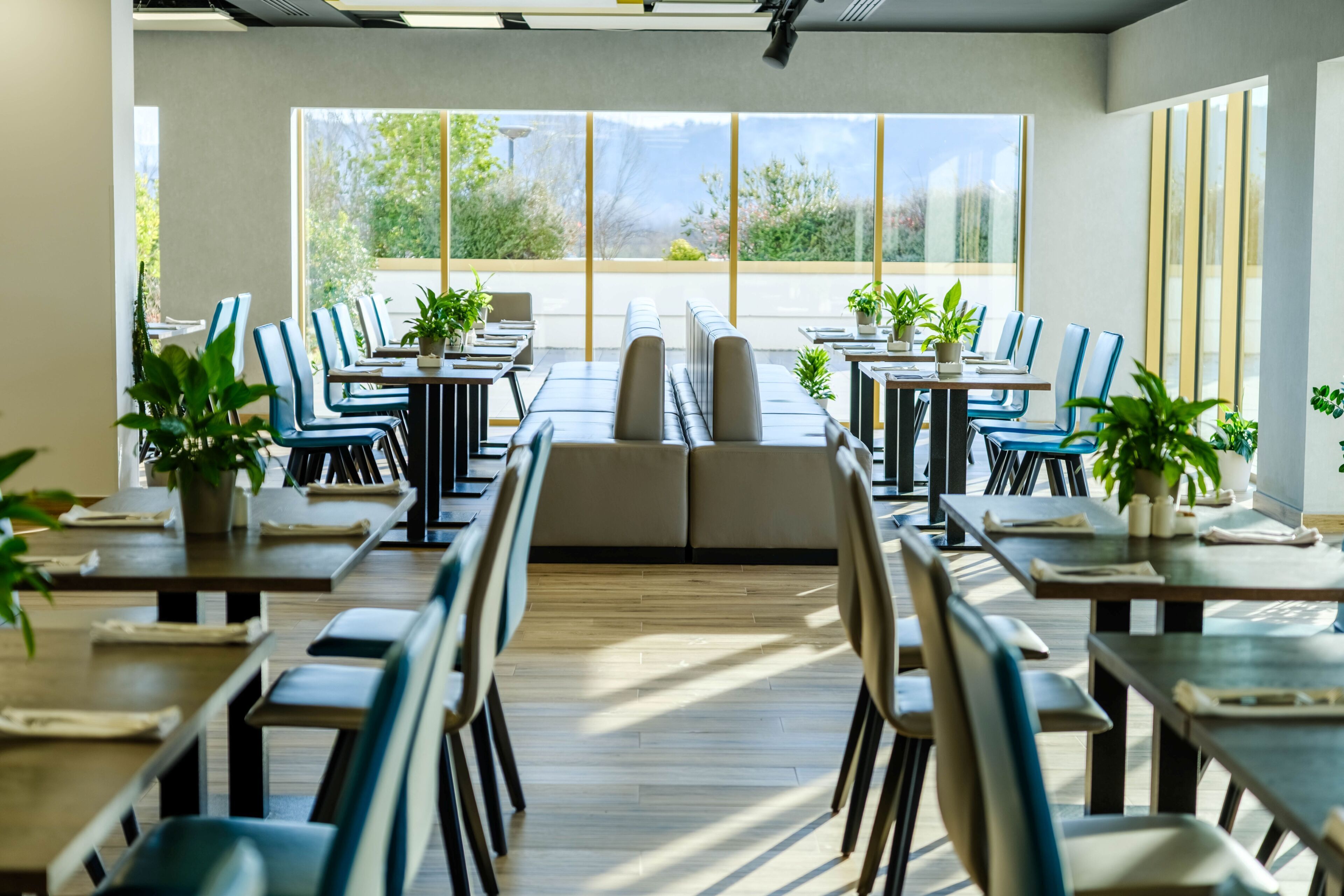 A spacious dining area with long tables, chairs, and potted plants.