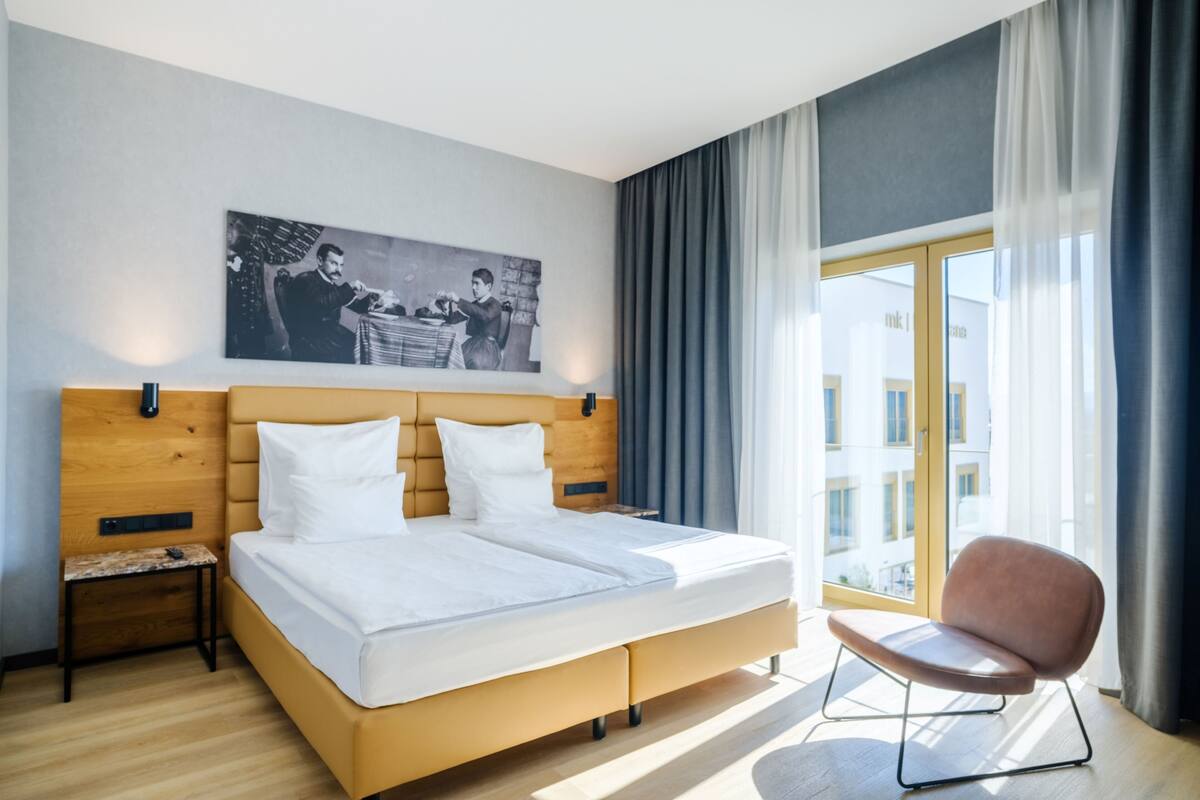 A modern hotel room with a large bed, a small side table, a chair, a large window with curtains, and a wall-mounted picture.