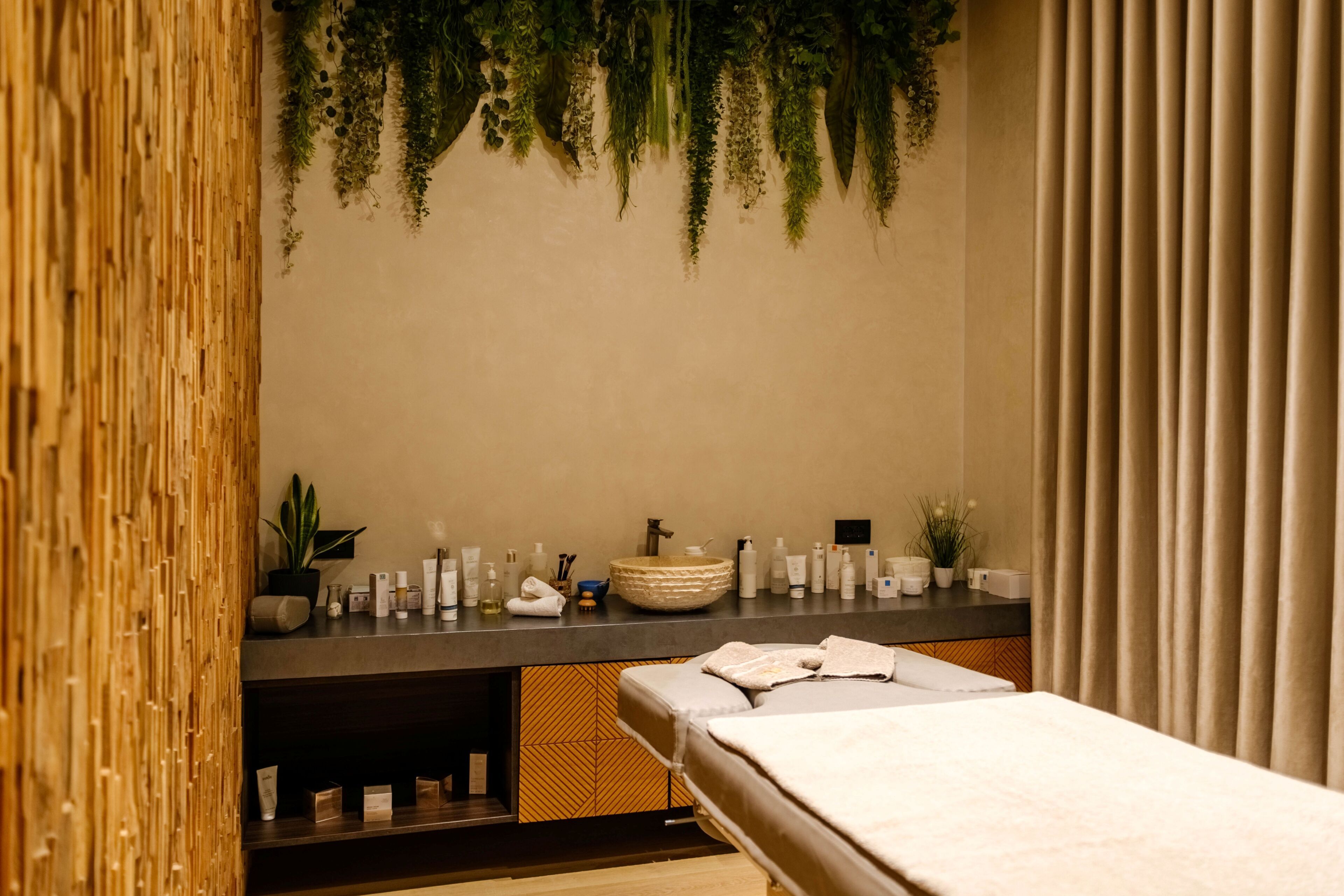 A spa room with a wooden wall, a large mirror, a sink with toiletries, a massage table, and hanging plants.