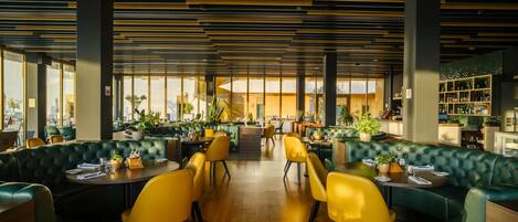 Un restaurant moderne avec des banquettes recouvertes d’un tissu vert, des chaises jaunes et un bar central.