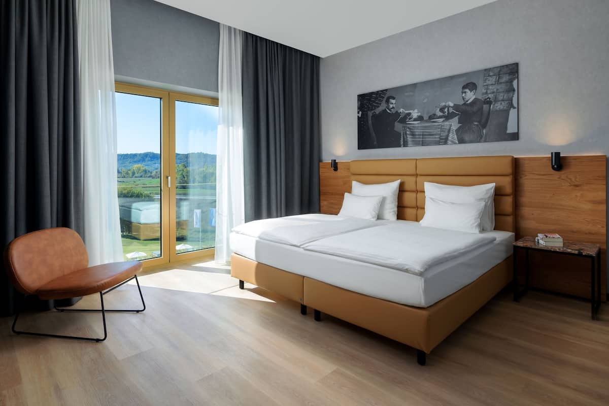 A modern hotel room with a large bed, a chair, a nightstand, and a painting on the wall.