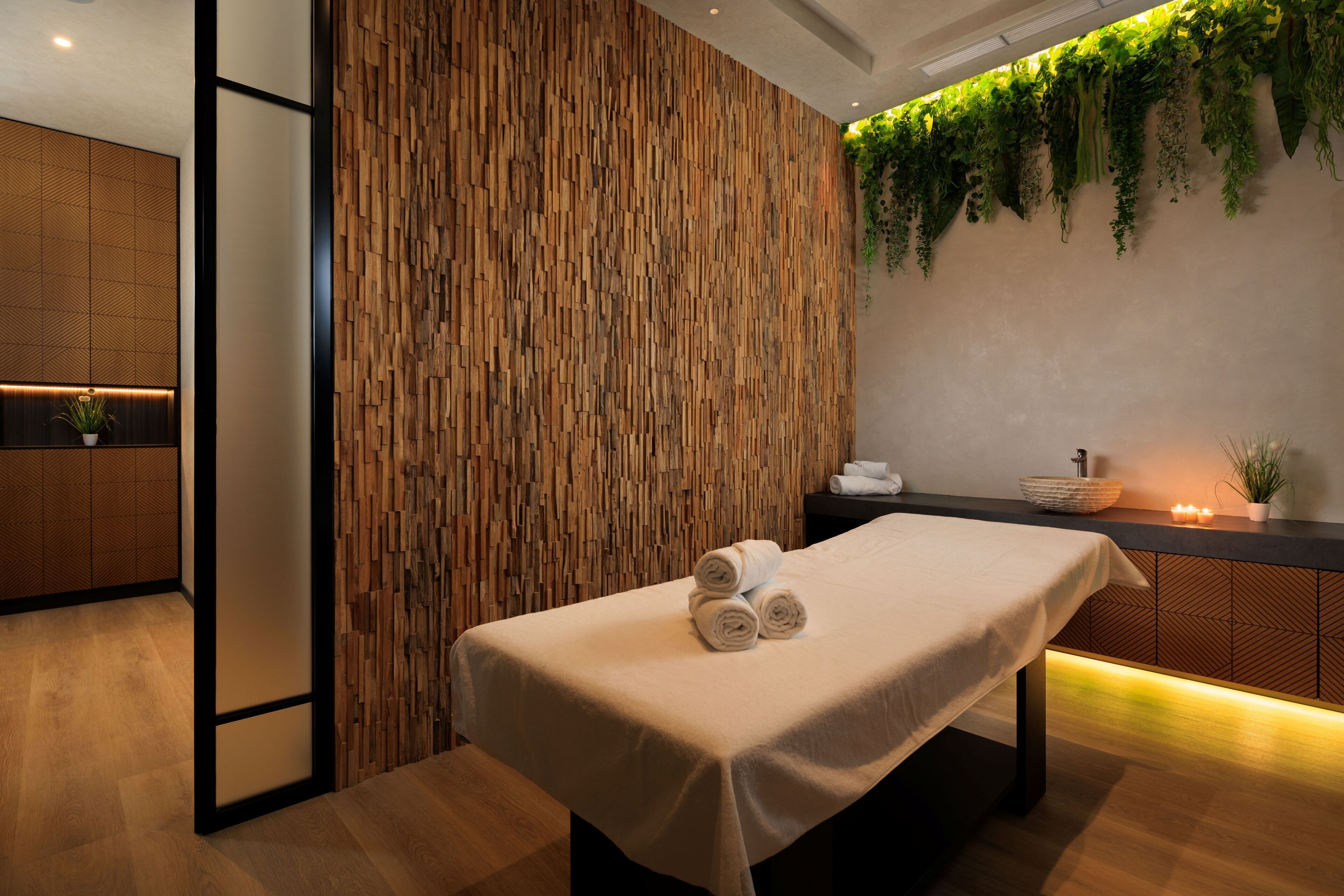 A spa room with a wooden wall, a massage table, a sink, and a plant.