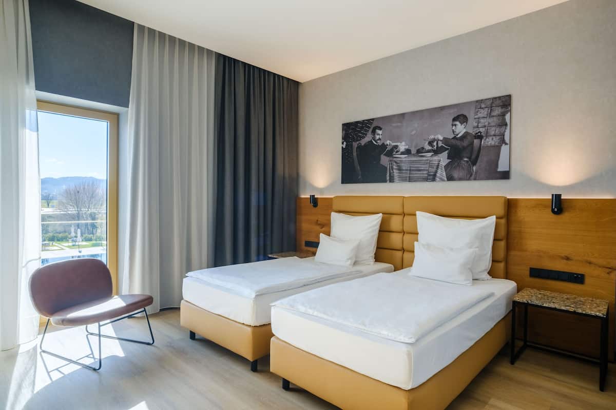A hotel room with two beds, a chair, a small table, a painting on the wall, and a window with curtains.