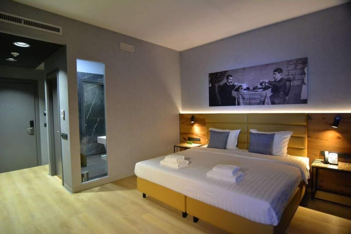 A modern hotel room with a large bed, a wooden headboard, a bedside table with a lamp, and a wall-mounted picture.