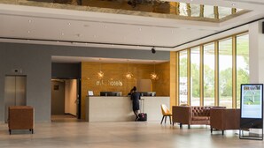 A modern hotel reception area with a receptionist, a reception desk, and a large window offering a view of the outdoors. - mk | hotel tirana (Preze)