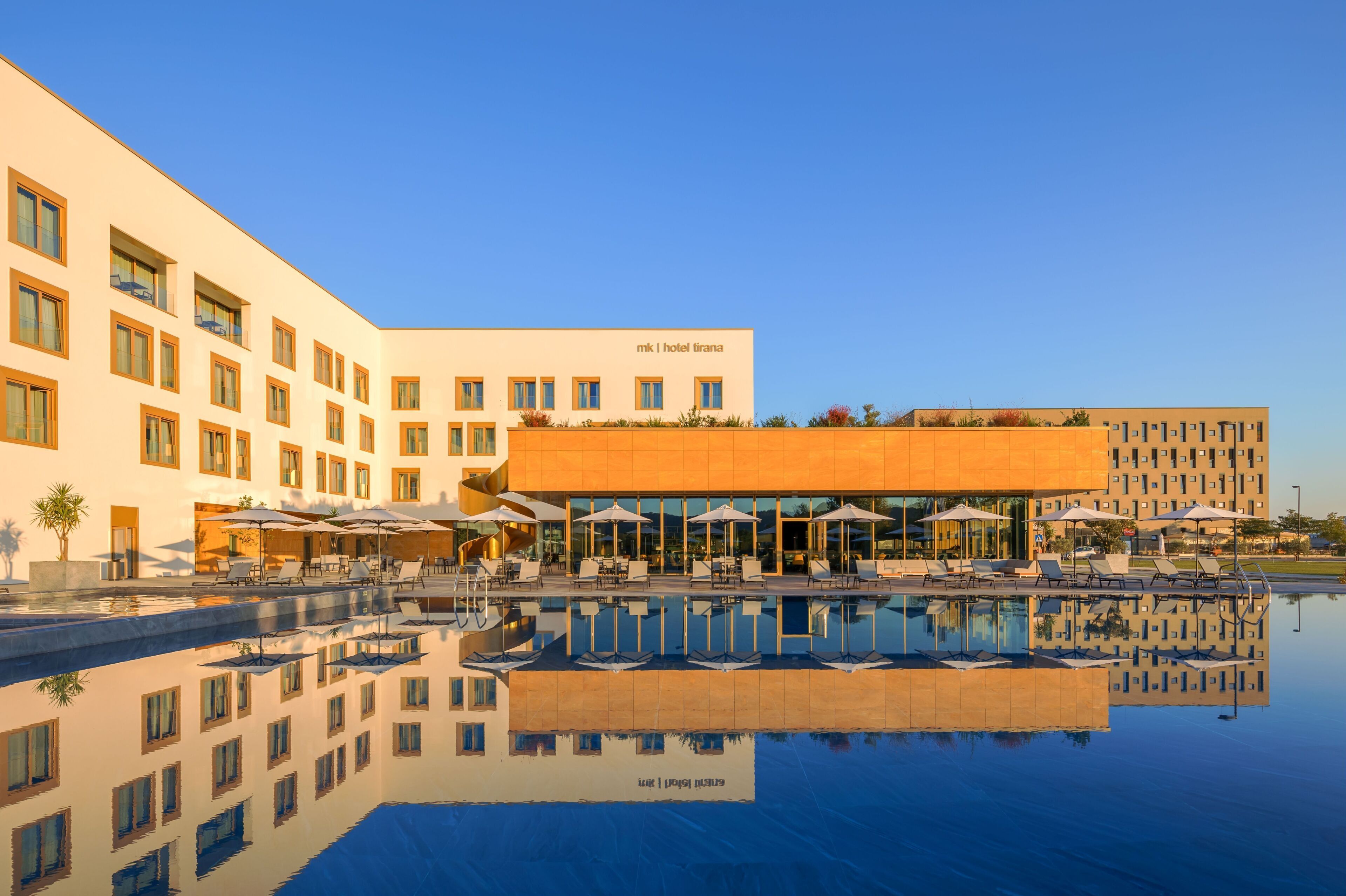 A hotel with a swimming pool, outdoor seating, and a clear sky.