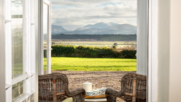 Una habitación con vistas a montañas a través de un ventanal, sillas de mimbre y una mesa con una taza y platillo.