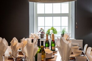 A dining table set with white napkins, wine bottles, and bowls, with a window and plants in the background. - Ty Fferam Bailey: A beautiful 7 bed holiday home in Anglesey (Bodorgan)