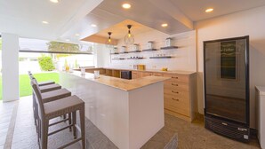 A modern kitchen with a central island, built-in wine cellar, and a view of a green outdoor area. - Cozy 3 Bedroom Pool Villa L70 (Hua Hin)