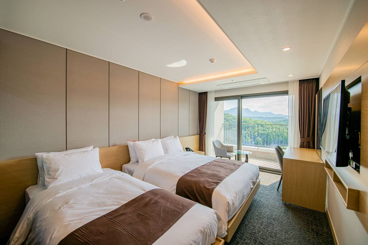 Habitación de hotel con dos camas, un escritorio, un televisor y un balcón con vista a la montaña.