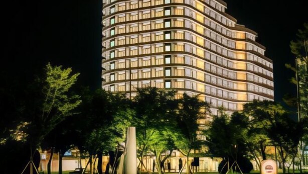 Een modern hotel met een gebogen gevel, verlichte ramen en een verzorgd buitengebied met bomen en een sculptuur.