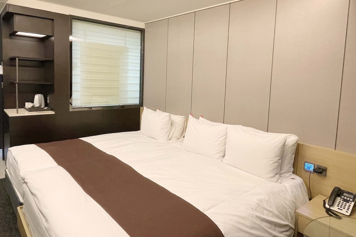 Habitación de hotel con una cama grande, un teléfono en la mesita de noche, un estante pequeño con artículos de aseo y una ventana con persianas.