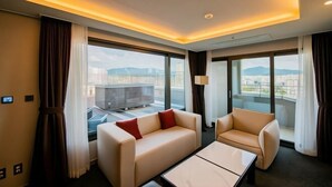 Een moderne hotelkamer met een bank, fauteuil en een klein tafeltje. Grote ramen bieden uitzicht op de stad en de bergen.