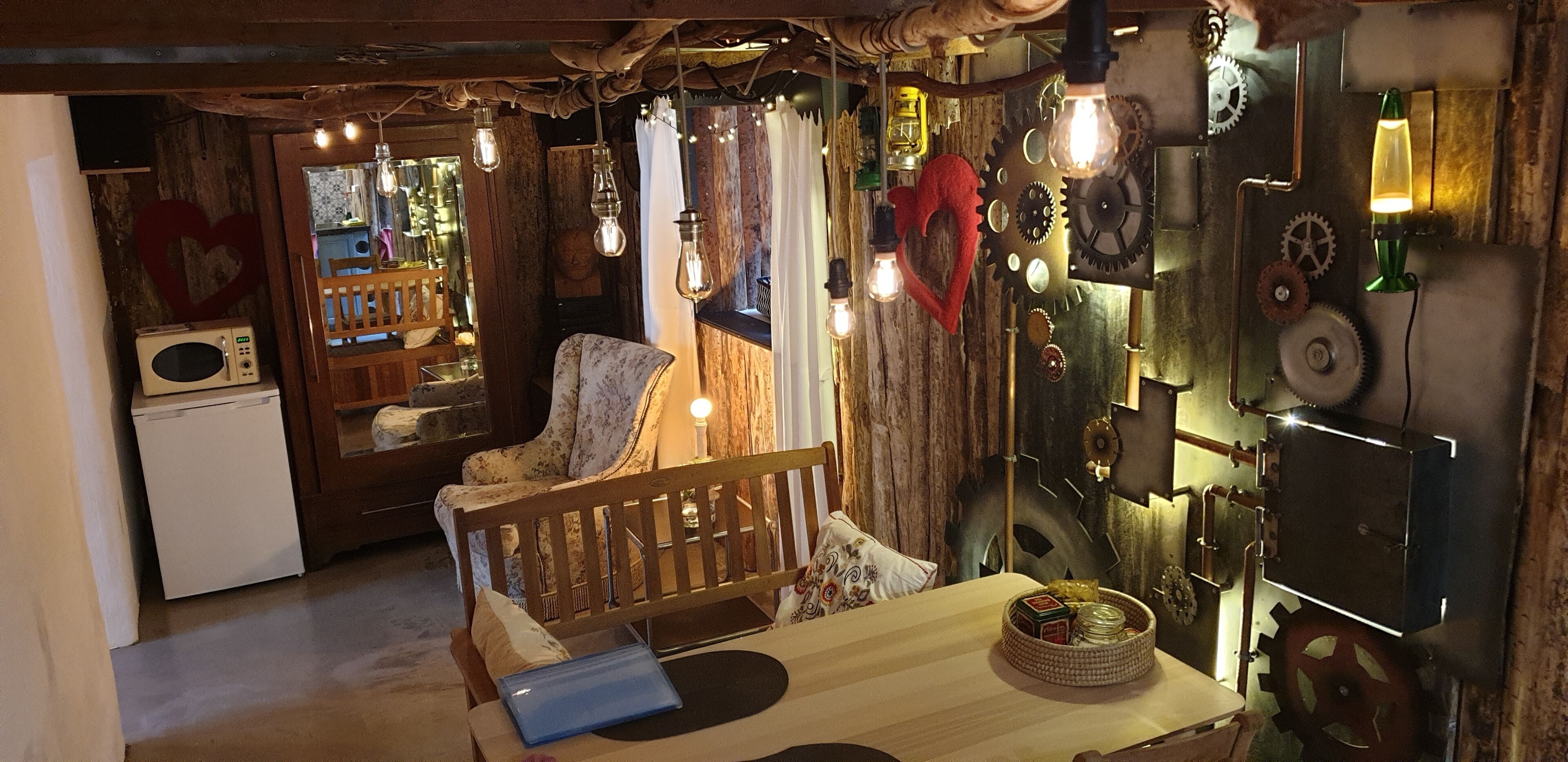 A cozy room with a wooden bench, a table with a laptop, and a wall adorned with gears and a lava lamp.