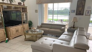 A living room with a beige sofa, a glass coffee table, a TV, and a view of a lake through large windows.