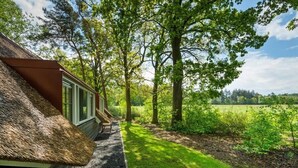 Un bâtiment à toit de chaume, doté d’une fenêtre et d’une porte, entouré d’arbres et bordé d’un champ d’herbe.