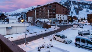 Une ville de montagne enneigée, avec un imposant bâtiment à plusieurs étages, une route bordée de véhicules garés et un ciel dégagé.