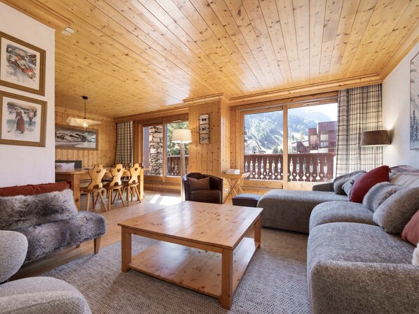 A cozy living room with wooden furniture, a stone fireplace, and large windows offering a view of the outdoors.