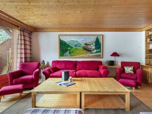 Uma sala de estar aconchegante com teto de madeira, um sofá vermelho, uma mesa de centro de madeira e uma pintura de paisagem montanhosa.