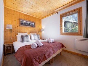 A bedroom with a wooden ceiling, a bed with a red bedspread, two bedside lamps, a painting on the wall, and a window with a view of trees.