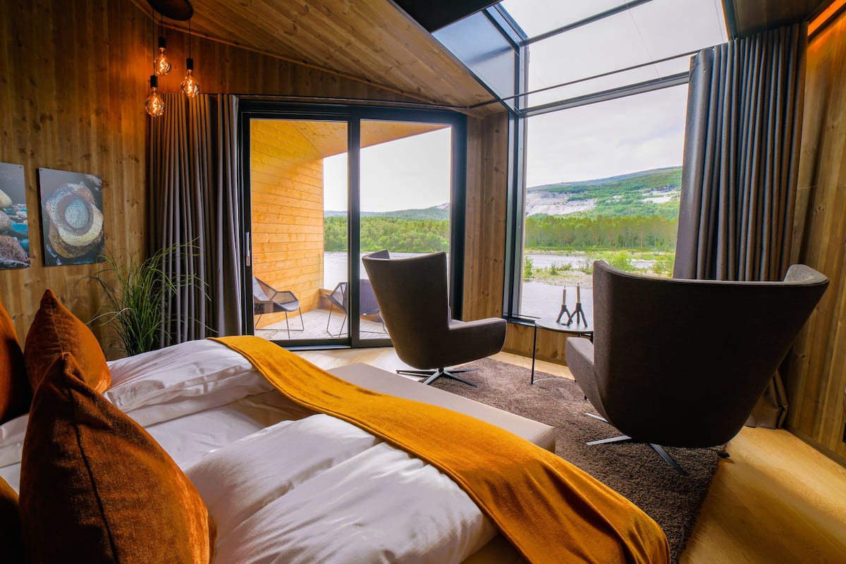 A modern bedroom with a large bed, a sofa, and a view of a natural landscape through large windows.