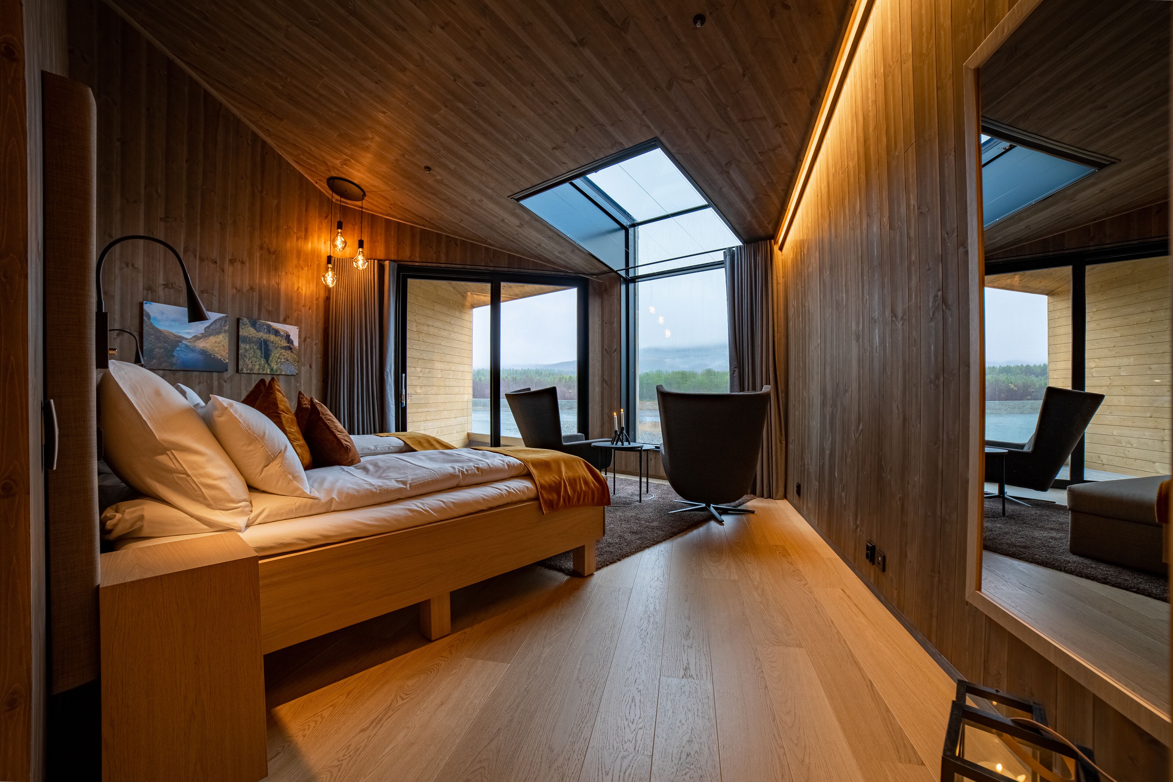 A modern bedroom with a wooden bed, large windows, and a view of the outdoors.