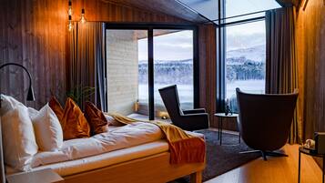 Ein modernes Schlafzimmer mit einem hölzernen Bett, einem Lounge-Sessel und einem großen Fenster mit Blick auf eine verschneite Landschaft.