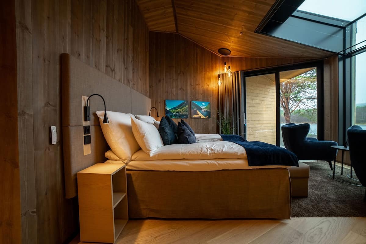 A modern bedroom with a wooden headboard, a bed with white bedding, a bedside table, and a view of the outdoors through large windows.