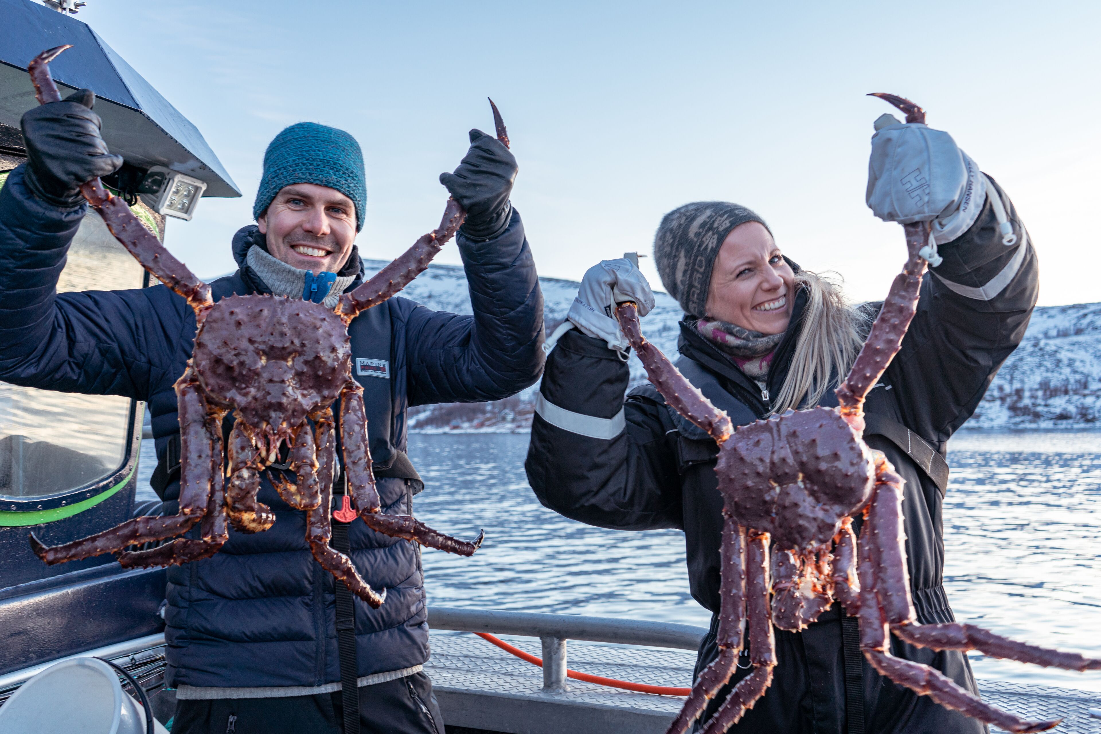 Two people holding large crab legs on a boat with a snowy landscape in the background.