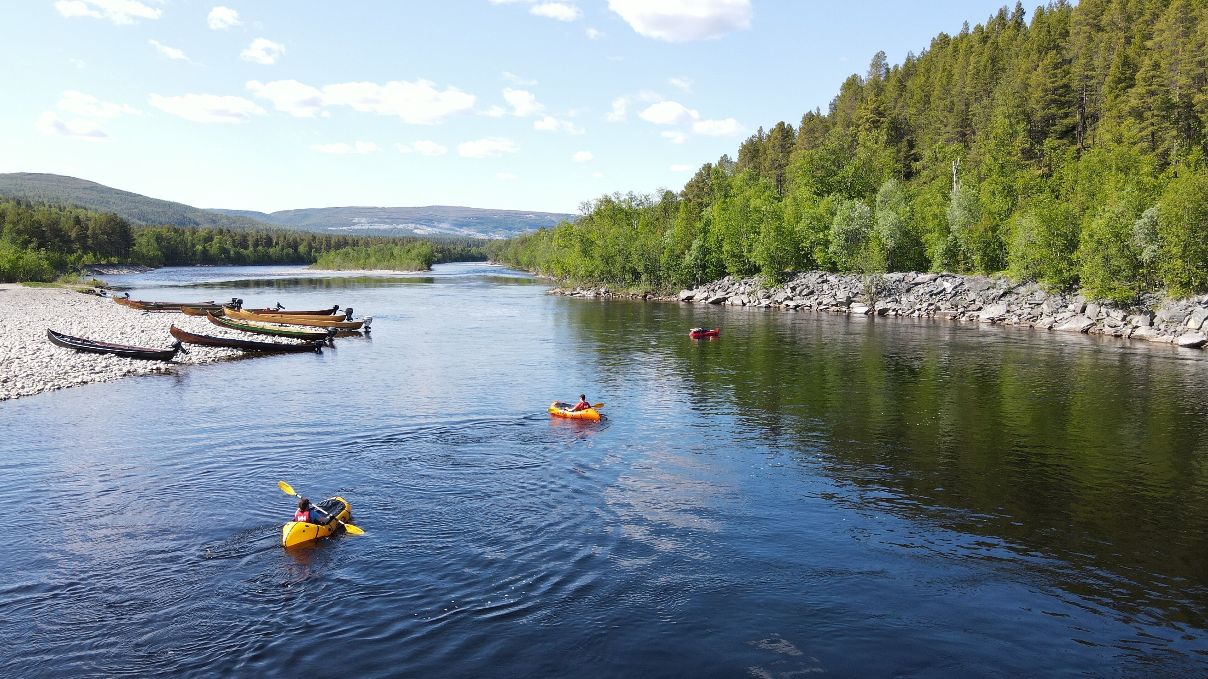 Ein Fluss mit Kajaks und Kanus am Ufer, umgeben von bewaldeten Hügeln.