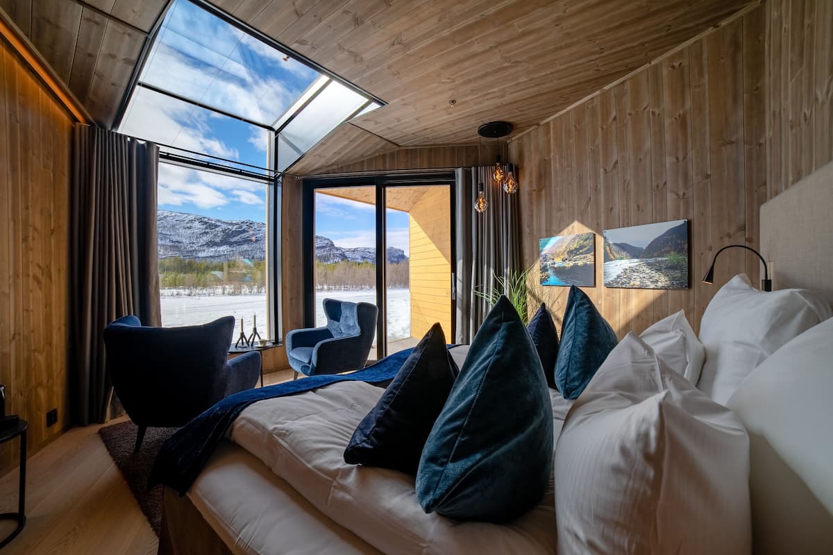 A modern room with a large bed, two armchairs, a skylight, and a view of mountains.