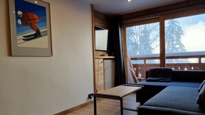 A cozy room with a wooden floor, a sofa, a small table, and a large window with a view of snow-covered trees.