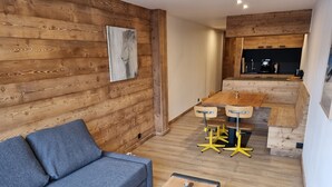 A cozy room with a wooden wall, a grey sofa, a small table, and a kitchen area with a wooden table and bar stools.
