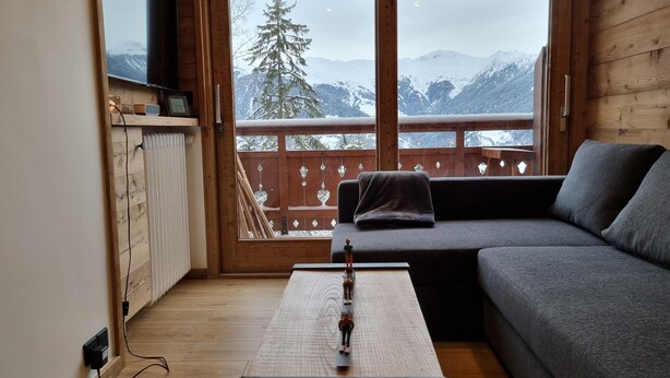 A cozy room with a wooden interior, a gray sofa, a small table with a decorative item, and a large window offering a snowy mountain view.