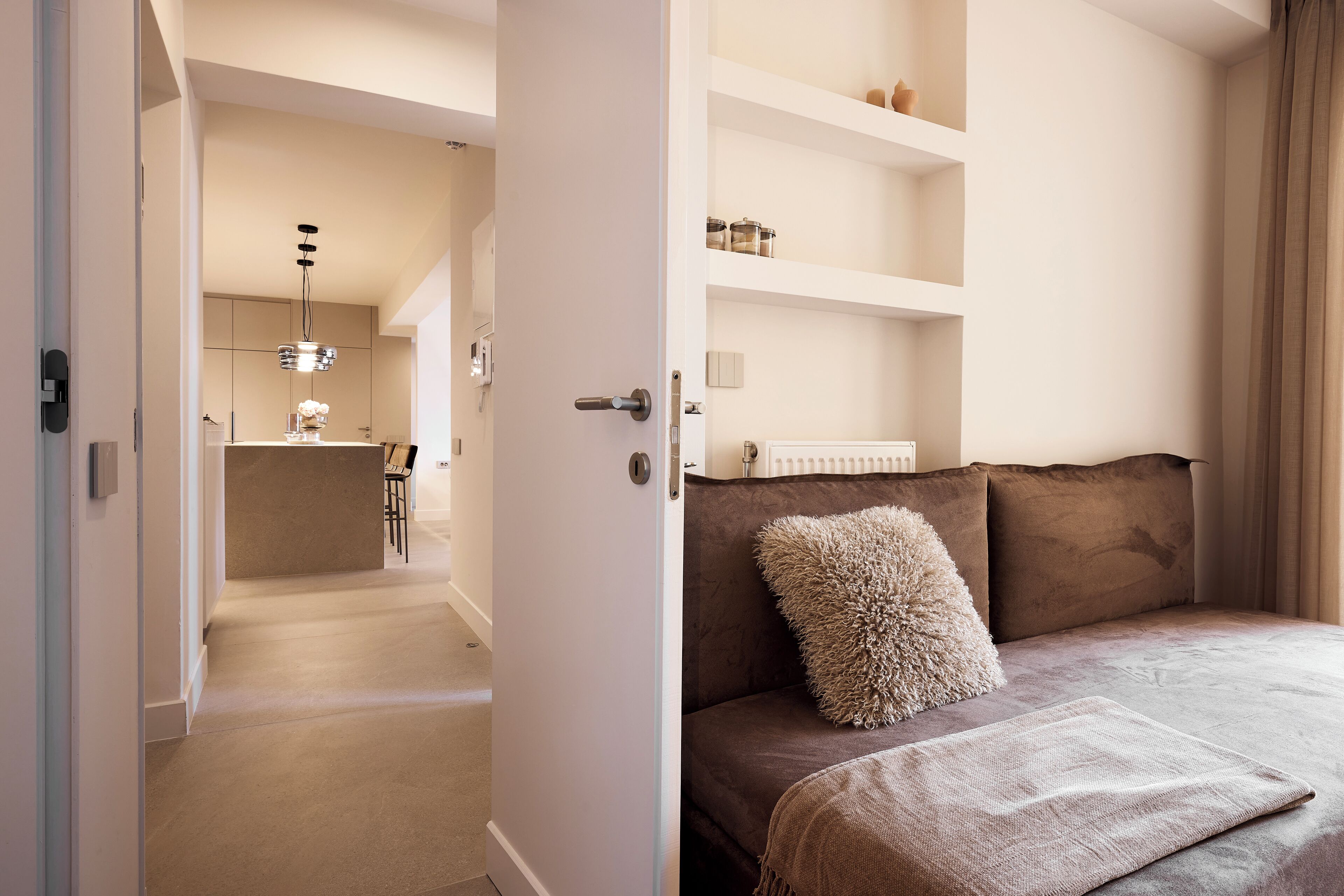 A modern hotel room with a sofa, a fluffy pillow, a radiator, and a kitchen area visible through an open door.