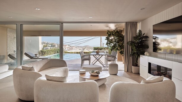 Ein modernes Wohnzimmer mit einem großen Panoramafenster, das auf einen Balkon mit Blick auf einen Pool führt. Es gibt einen Essbereich mit Tisch und Stühlen sowie einen Kamin und einen Fernseher.