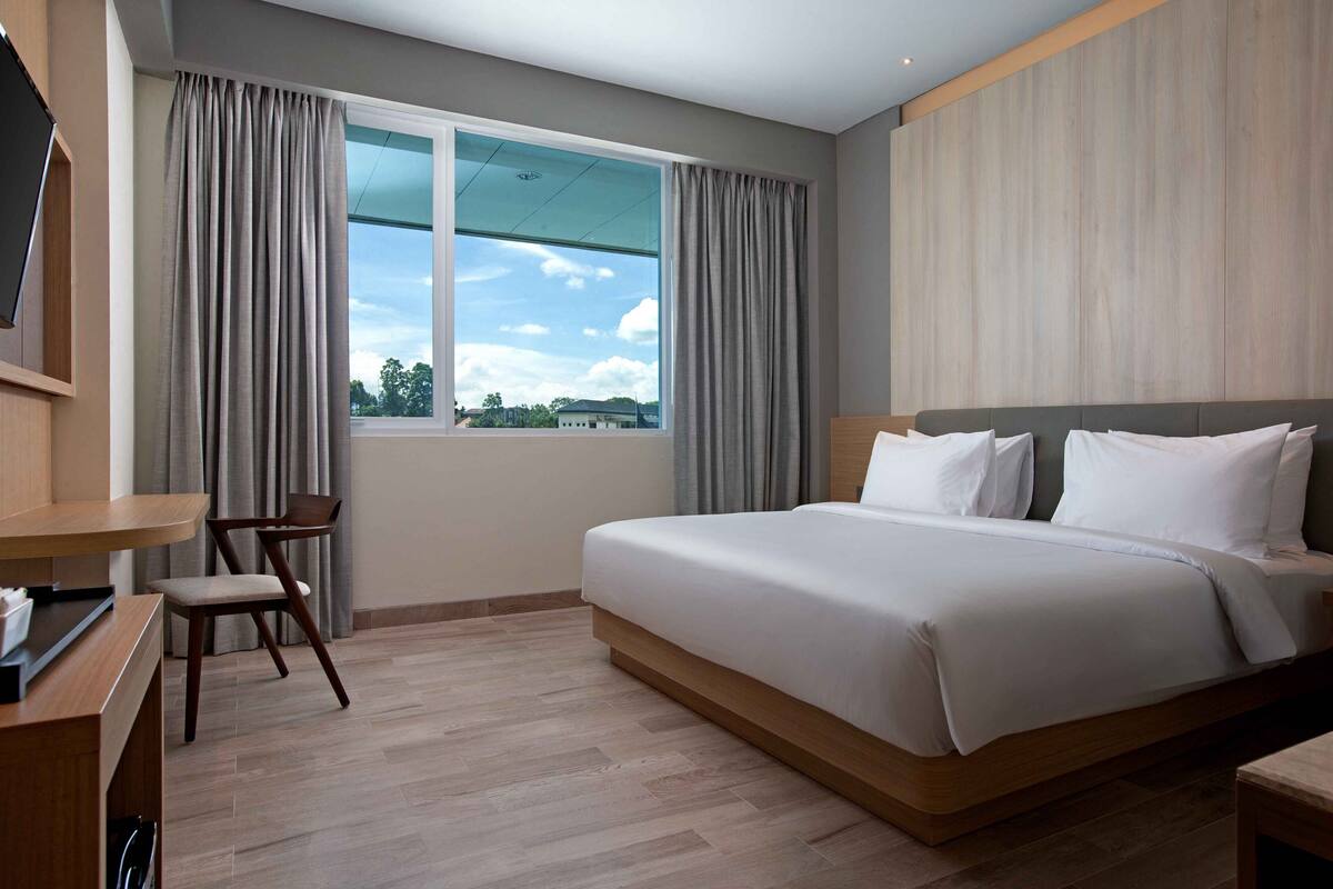 A modern hotel room with a large bed, a desk with a chair, and a window with curtains.