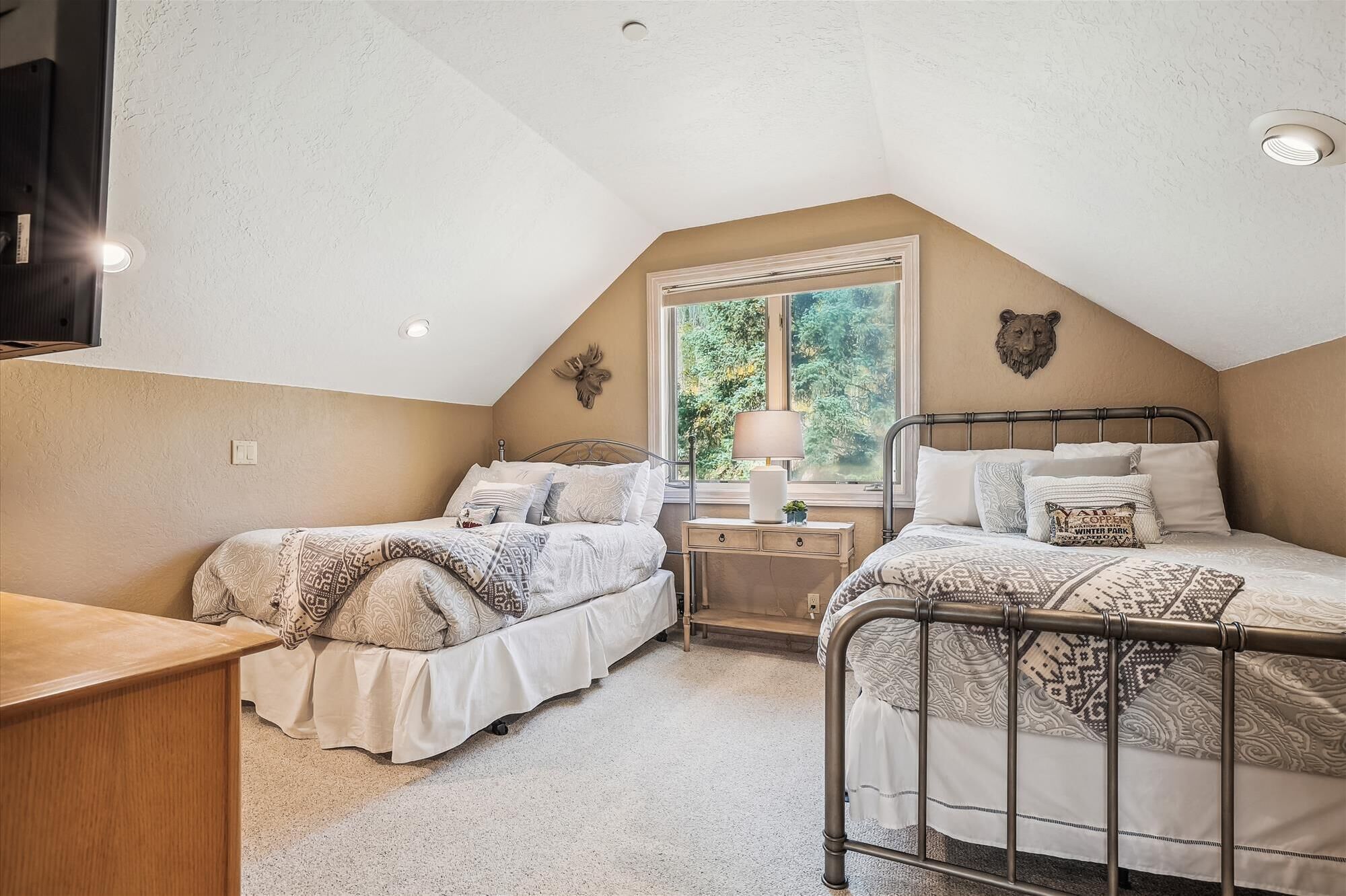 A bedroom with two single beds, a wooden desk, a window with a view of greenery, and a bear head decoration on the wall.