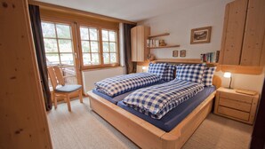 A bedroom with a wooden bed, a chair, a nightstand, and a window with a view of greenery. - Haus Lizi - Apartment 8 (Zermatt)