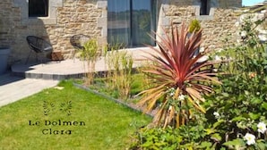 Une maison en pierre dotée d’une porte en verre moderne, d’un petit espace extérieur aménagé avec des chaises et d’un jardin bien entretenu où poussent différentes espèces de plantes.