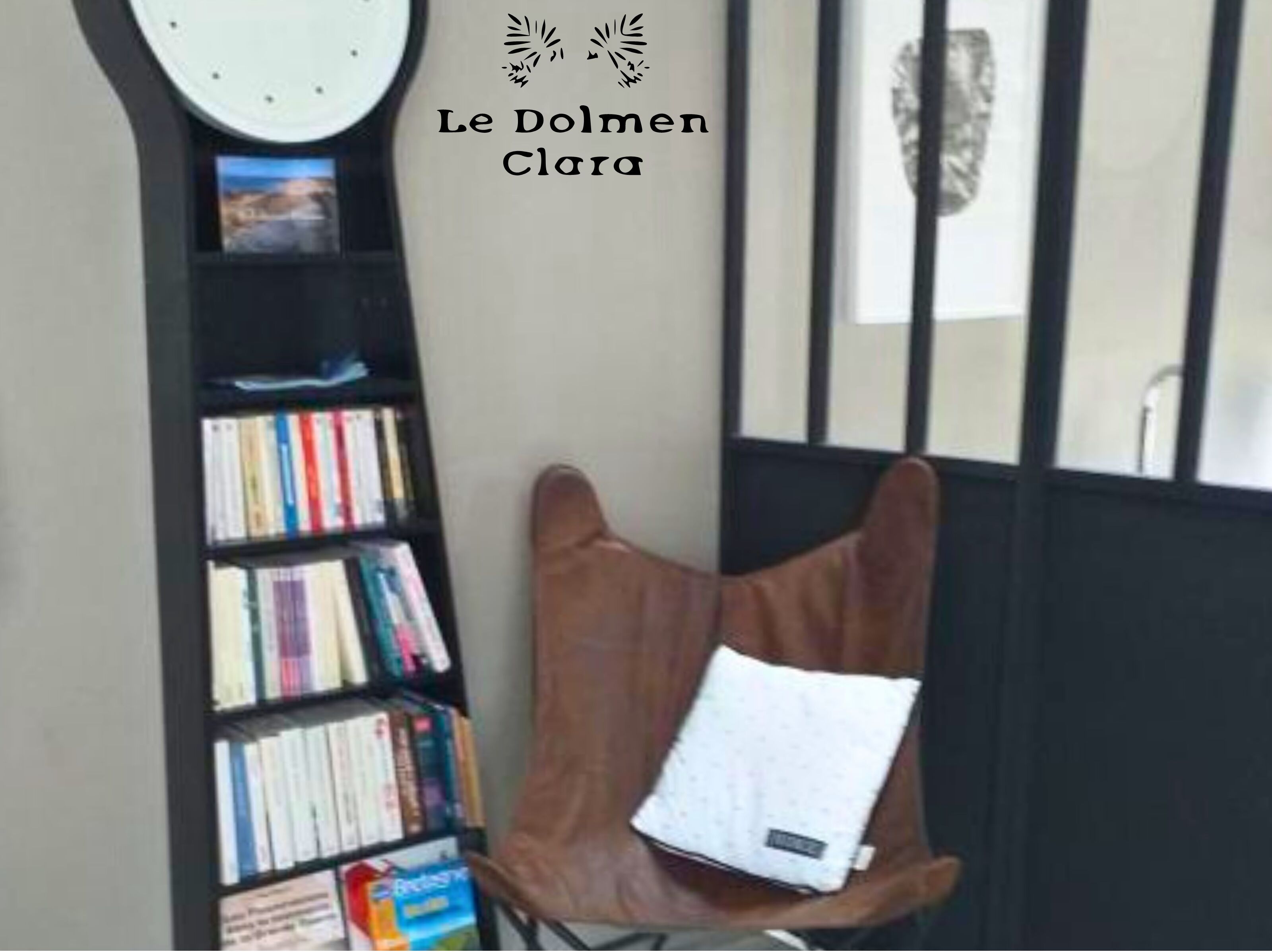 Una habitación con una estantería llena de libros, un sillón marrón con un cojín y un reloj de pared.