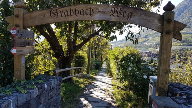 A wooden signpost with directions to various locations, a stone pathway, and a view of a valley with buildings. - Spacious Apartment “Lafreid Algund” near Merano with Mountain View, Wi-Fi & Garden (Algund)
