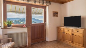 A room with a wooden door, a radiator, a chair, a TV on a wooden cabinet, and a window with a view of mountains.